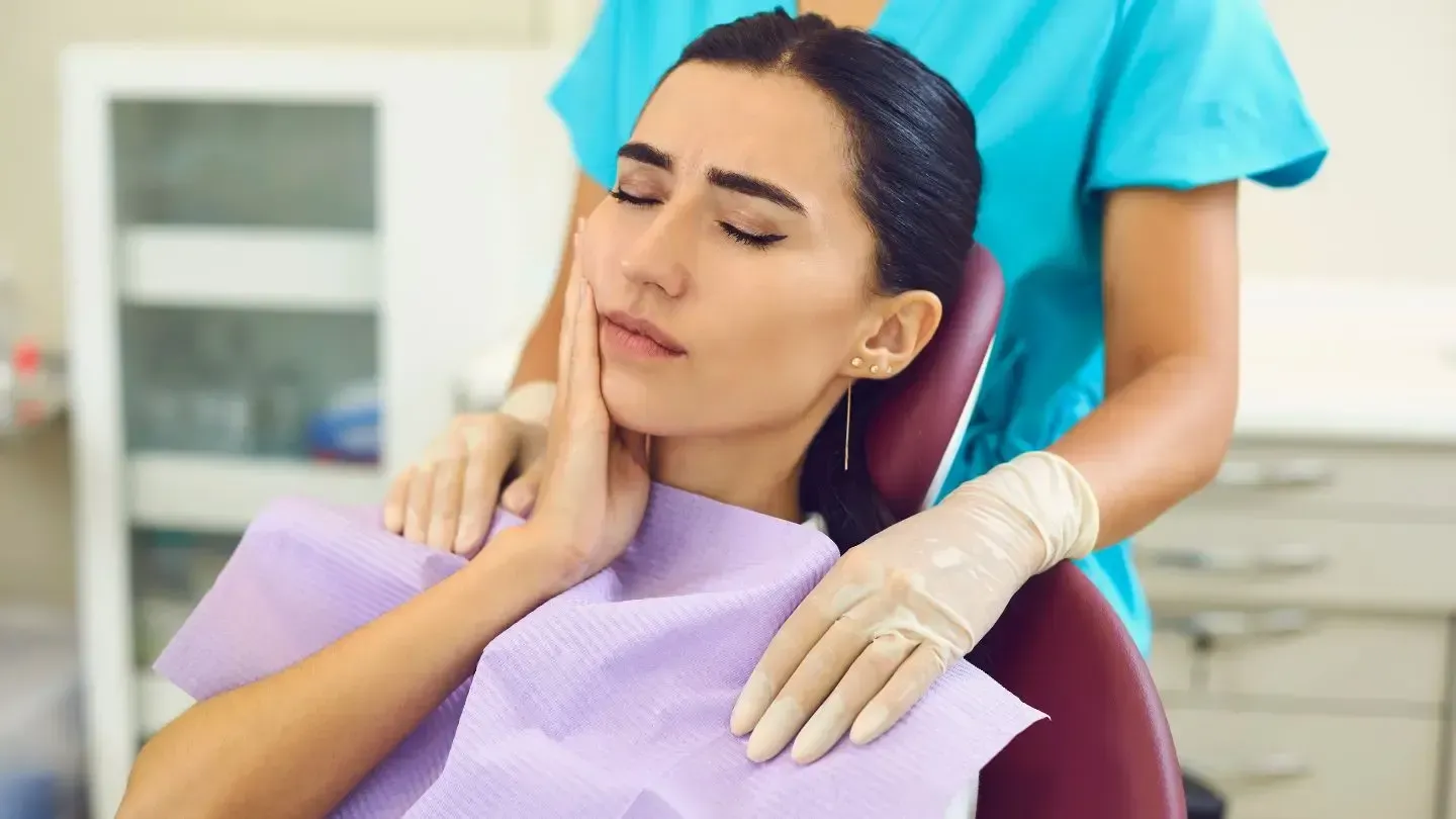 Woman in dental chair holding jaw, appearing in pain, dental assistant nearby.