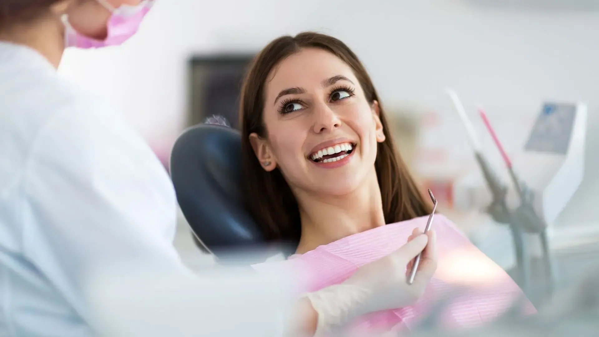 Woman smiling at a dentist in a dental office. The dentist wears a mask and gloves.
