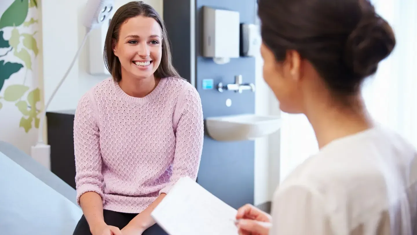 A woman in a pink sweater smiles at a healthcare provider in an exam room. They are discussing a document.