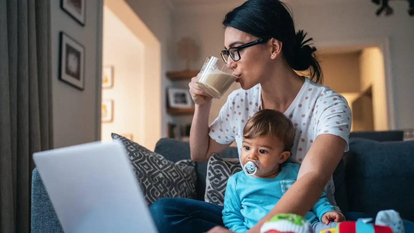 Woman with dark hair and glasses sips a drink while working on a laptop. A baby with a pacifier sits in her lap.