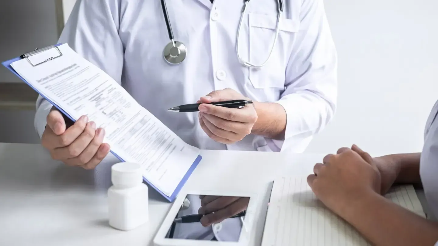 Doctor in white coat pointing at a medical form on a clipboard, seated across from a patient at a desk.