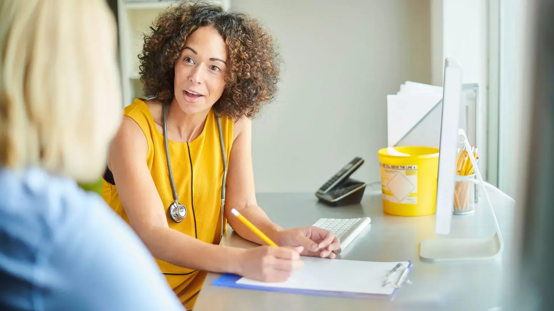 Doctor in yellow dress with stethoscope, talking to patient at desk, writing notes.