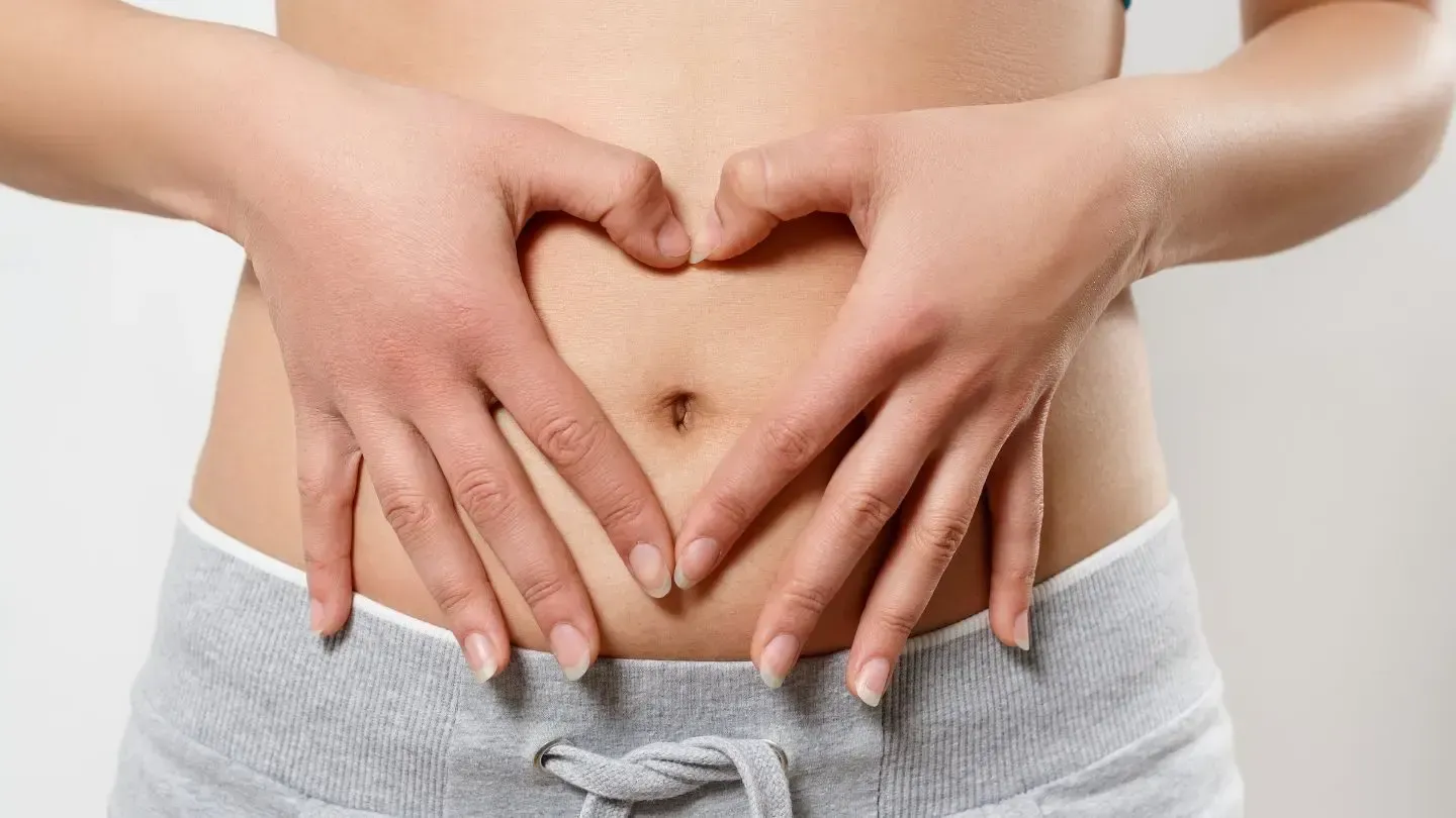 Hands forming a heart over a person’s abdomen, wearing gray sweatpants, white background.