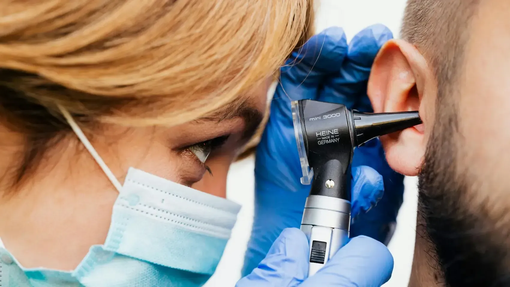 Doctor examining a patient's ear with an otoscope. Blue gloves and surgical mask visible.