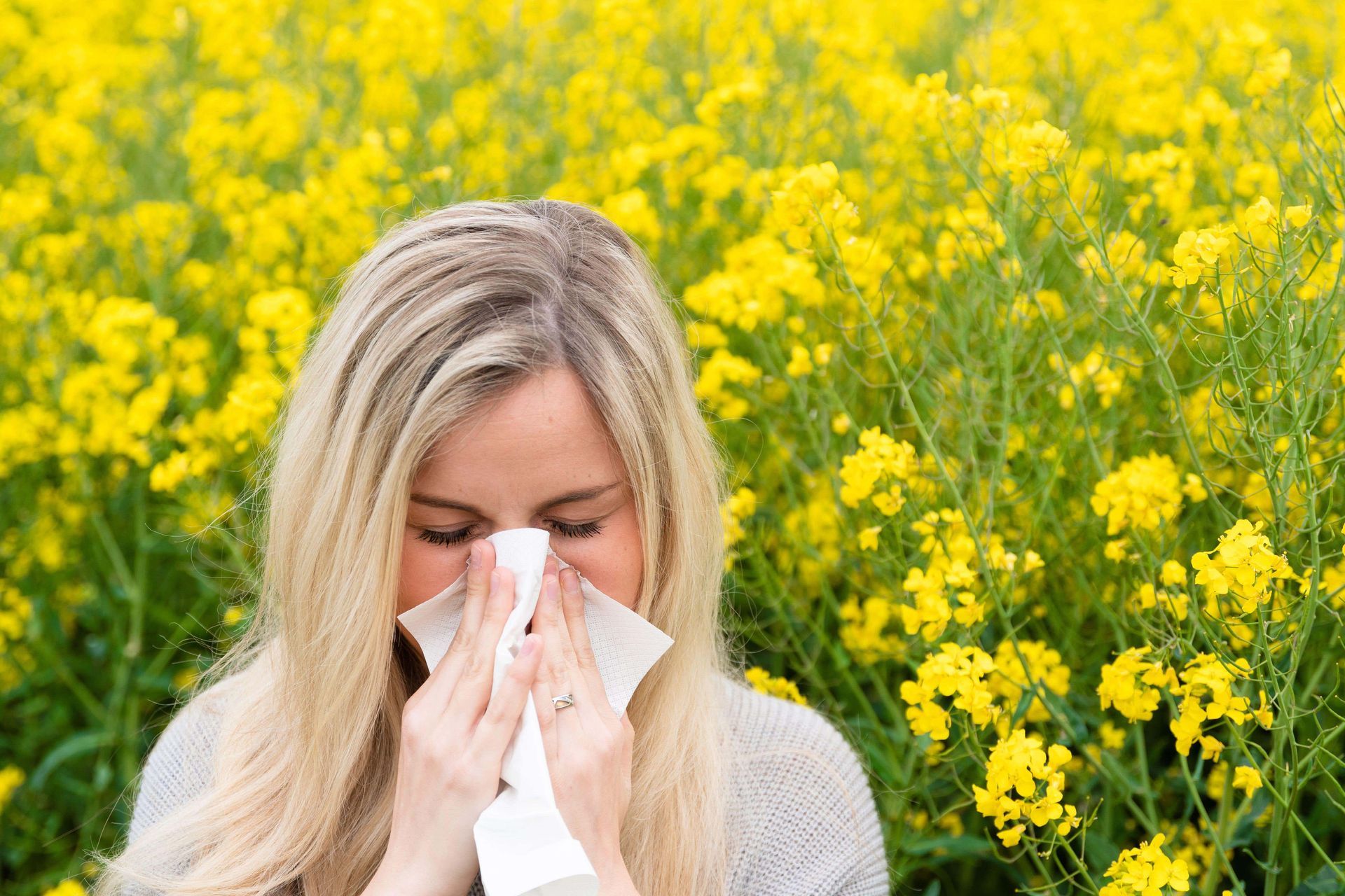 Woman sneezing into tissue, surrounded by yellow flowers in a field.