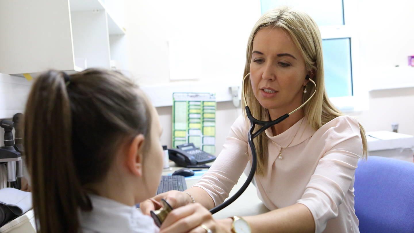 A female doctor using a stethoscope to examine a young girl's chest in a medical office.