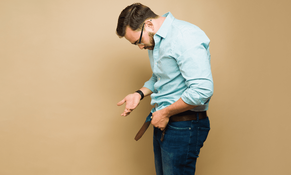 Man with glasses looking down, unbuckling belt, against a tan background.