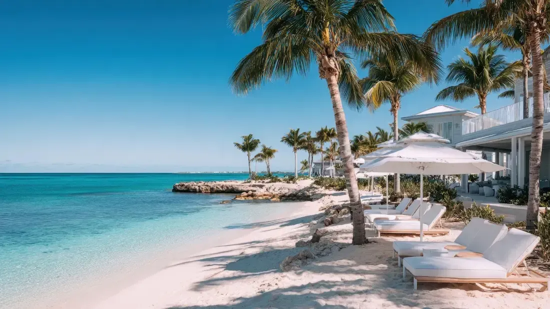 Beach scene with white sand, turquoise water, palm trees, lounge chairs, and buildings under a clear blue sky.