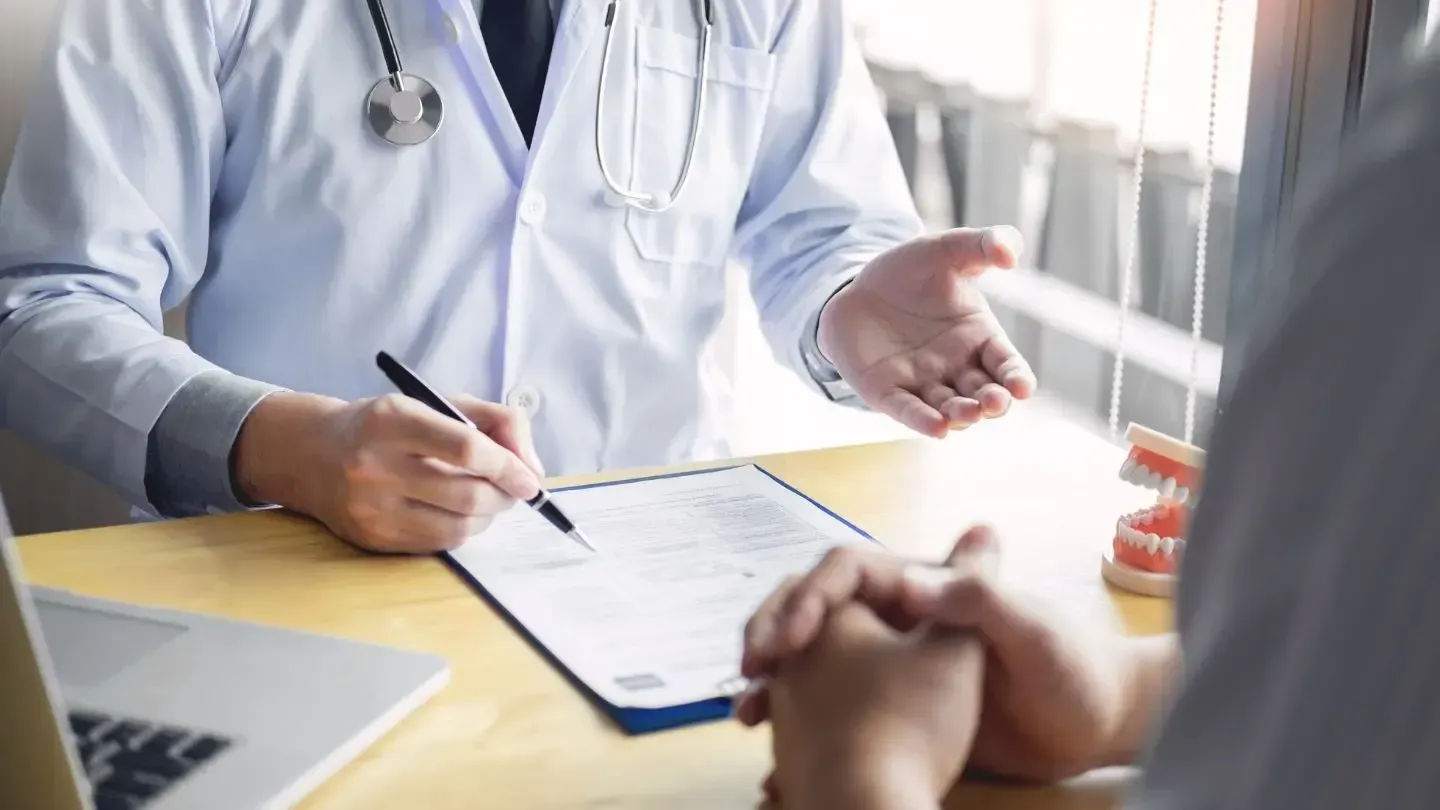 Dentist in a white coat explaining forms to a patient, teeth model on desk.