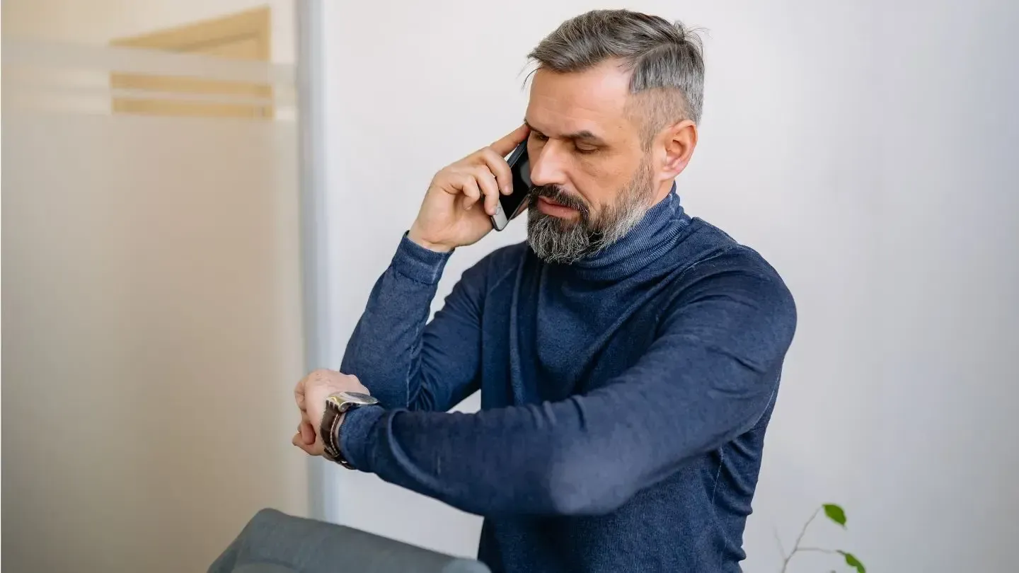 Man in blue turtleneck on phone, checking his watch, indoor setting.