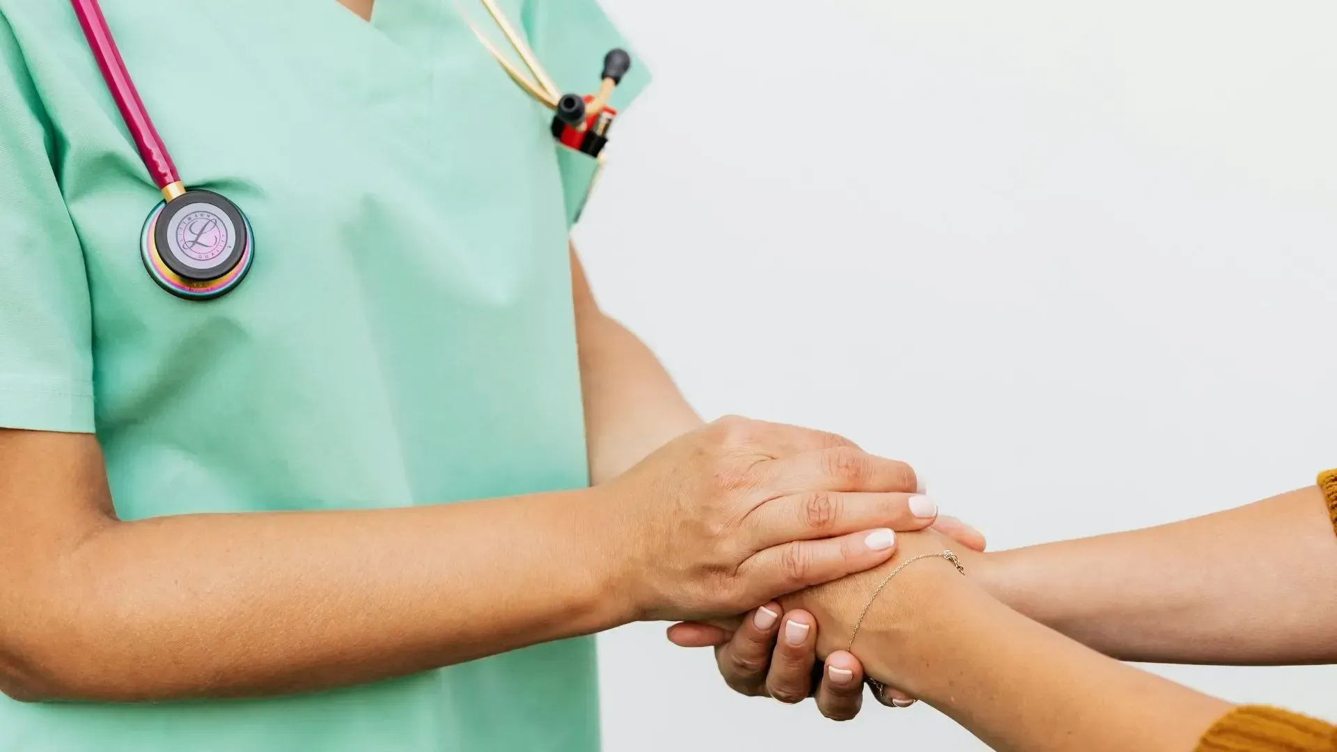 A healthcare professional in light green scrubs holding a patient's hand in a gesture of support.