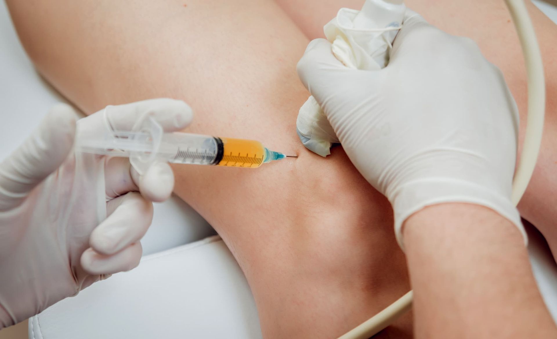 Medical professional injecting substance into knee; sterile gloves, syringe, ultrasound probe present.