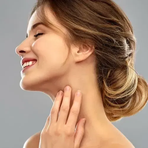 Woman smiling, touching her neck. Light skin, brown hair in bun, gray background.