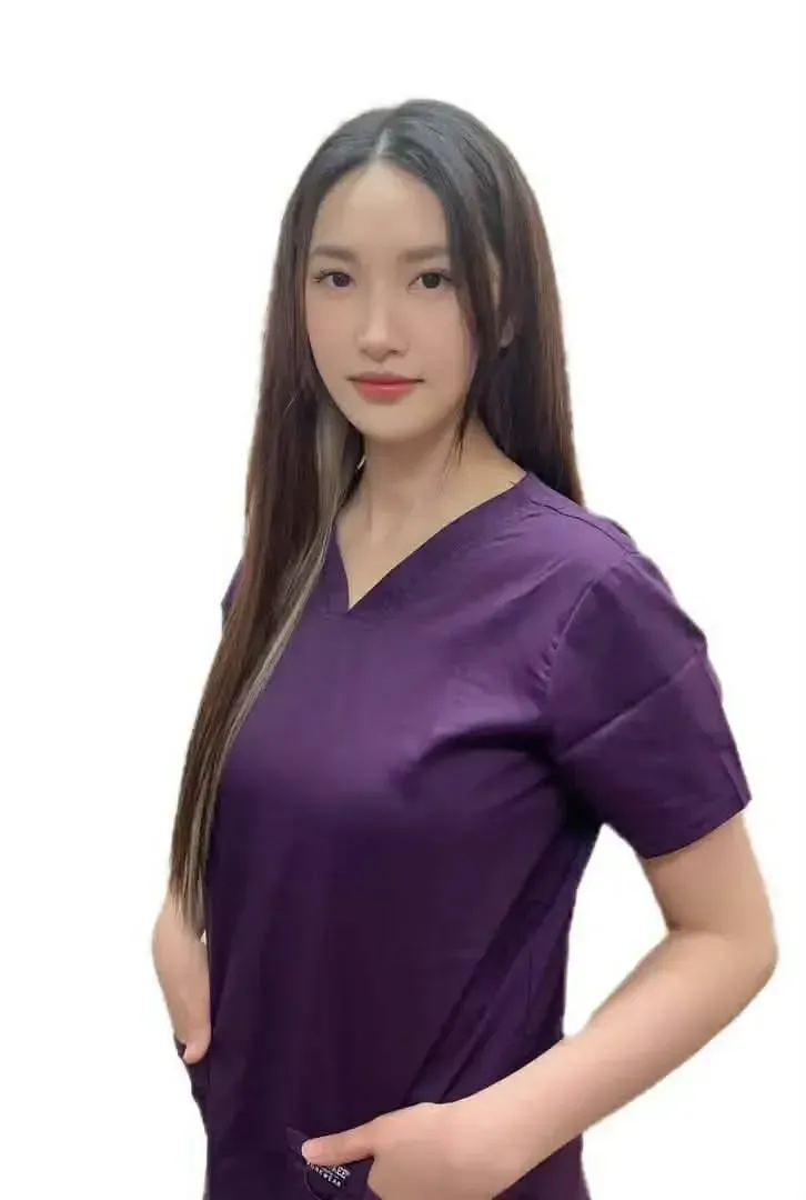 A person with long dark hair, wearing a purple V-neck scrub top, standing with hands in their pockets against a white.