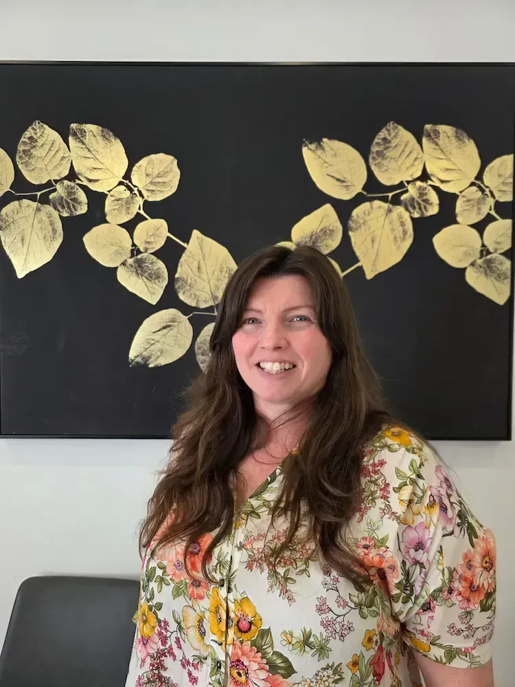 Woman smiling indoors in front of a black wall with gold leaf artwork, wearing a floral blouse.