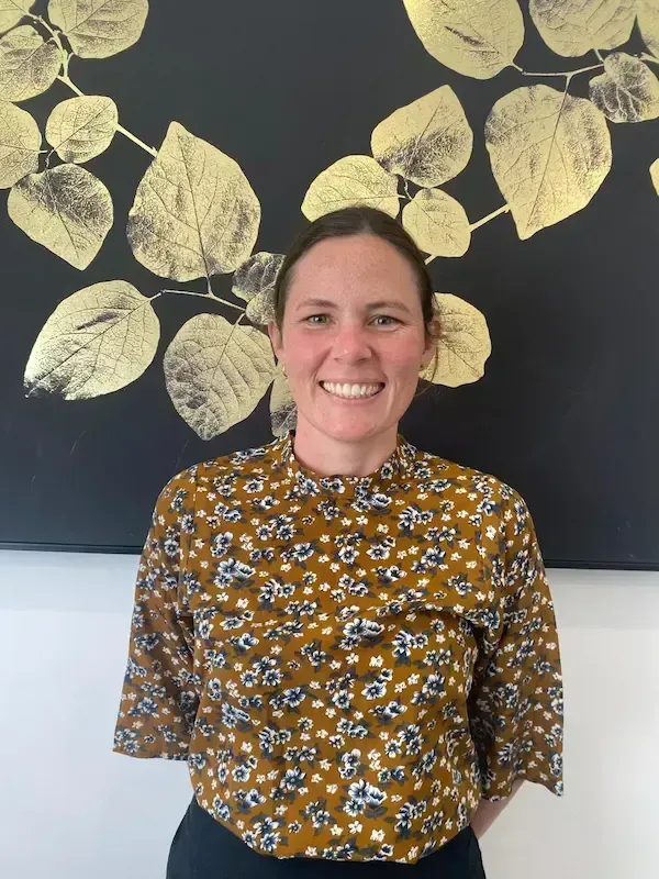 Smiling person in a mustard floral top standing before a black wall with gold leaf artwork.