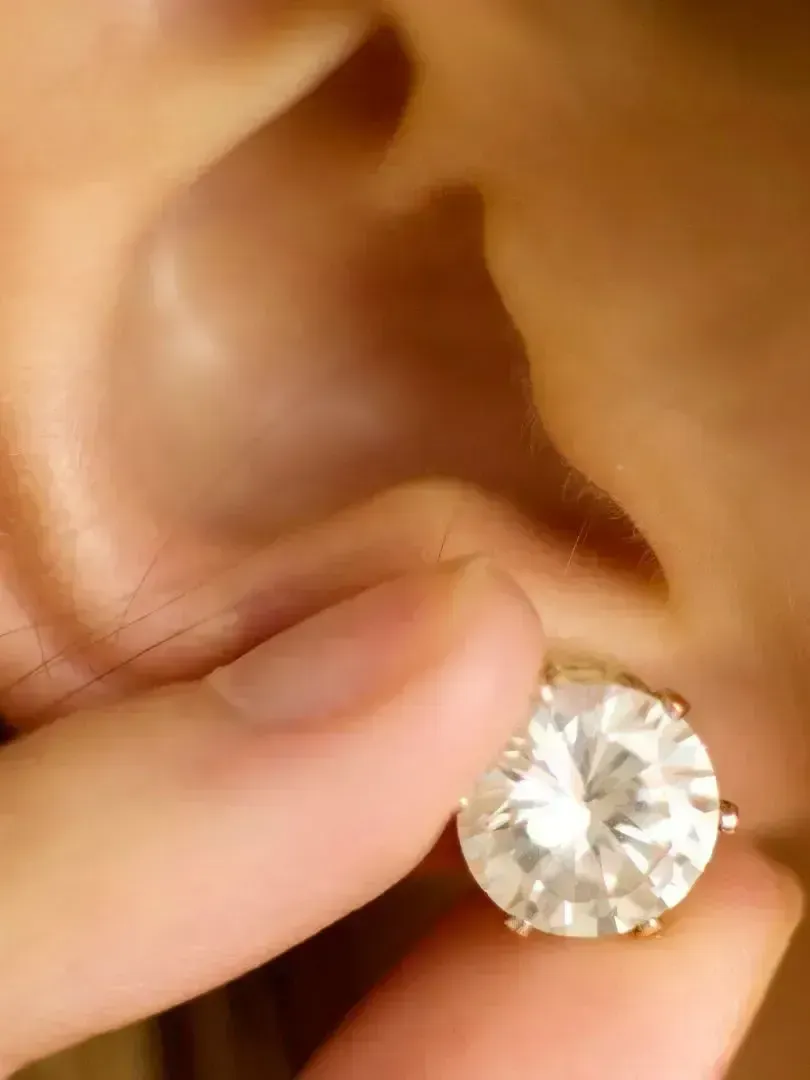 Hand holding a large, round, white gemstone earring near an ear.