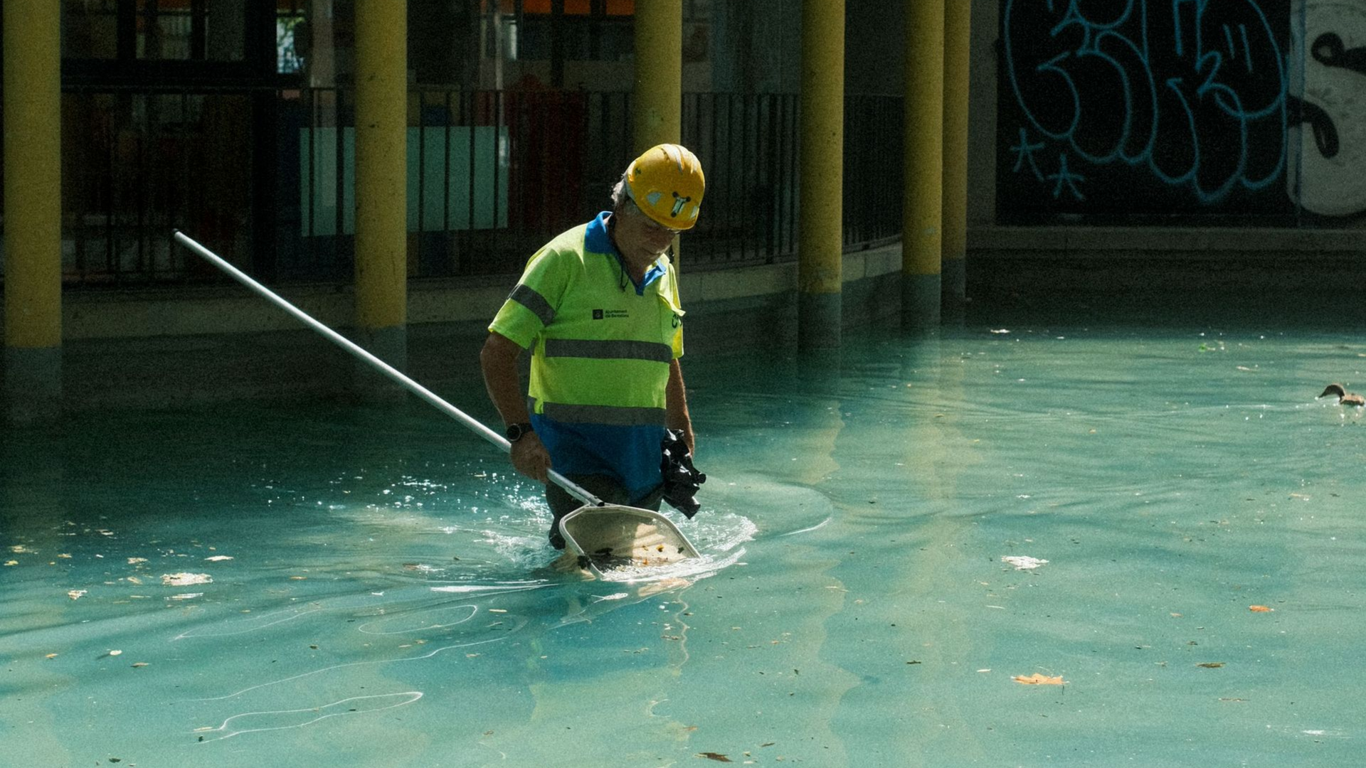 Worker in yellow hard hat standing in flooded water, using a hose or tool in an industrial area.
