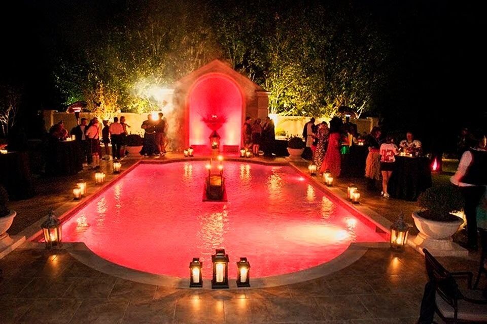 Night-lit pink pool with lanterns and guests gathered around a glowing arched fountain.