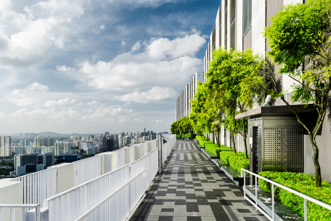 Rooftop walkway beside modern apartments, with white railings, green vines, and a city skyline under a cloudy sky