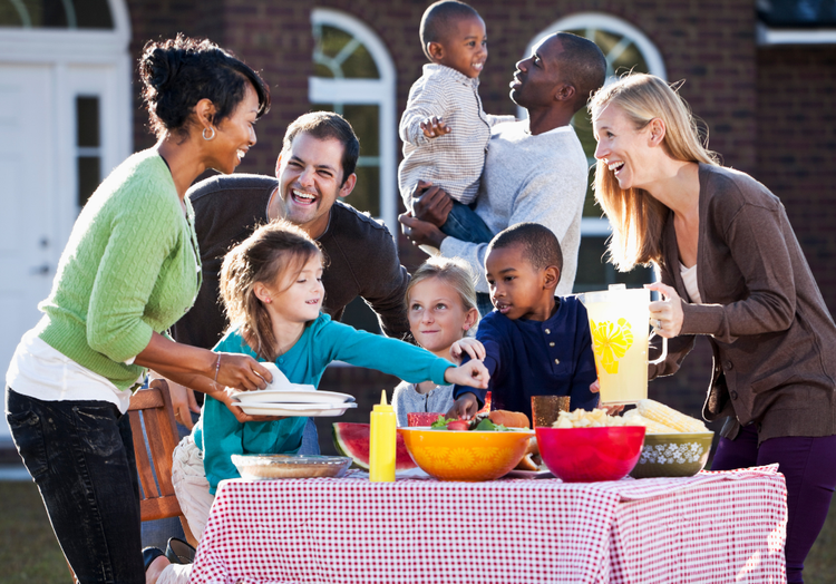 People sharing food and drinks at an outdoor picnic table, smiling and chatting in a backyard.