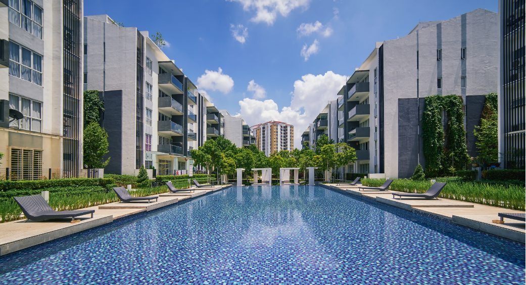 Modern apartment complex with a long blue swimming pool and landscaped walkways under a partly cloudy sky