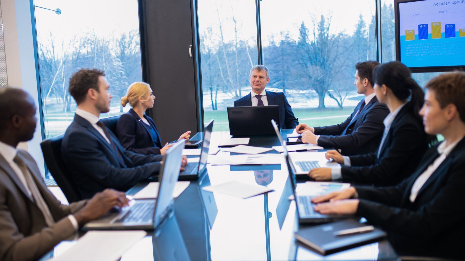 Business meeting around a conference table, with a presenter speaking at the front and a chart on sc