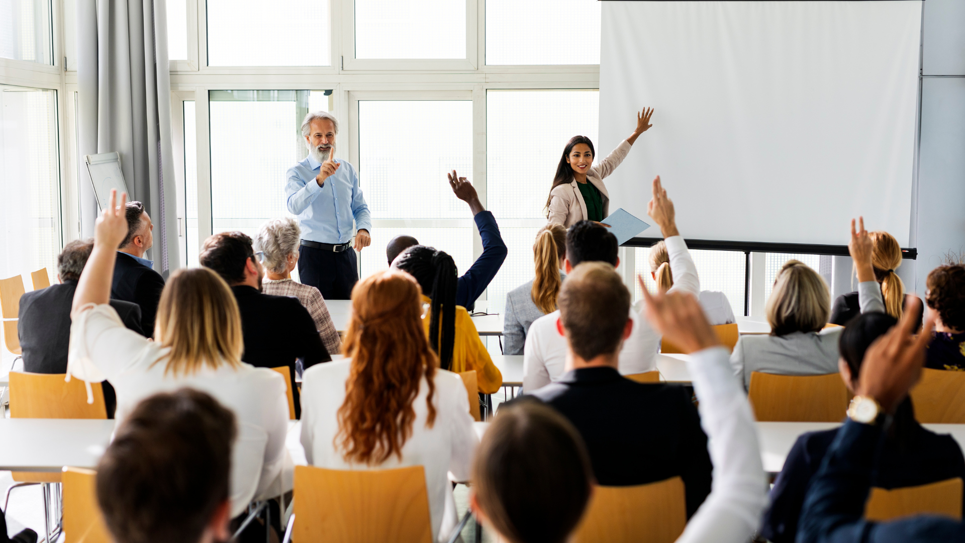 Audience in a bright conference room raising hands during a presentation on stage