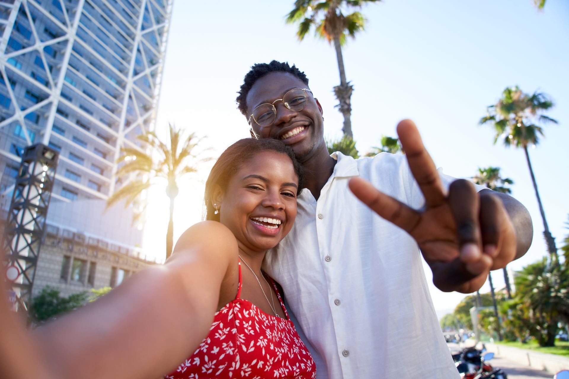 Two people smiling for a selfie beside palm trees and a tall building in bright sunlight.
