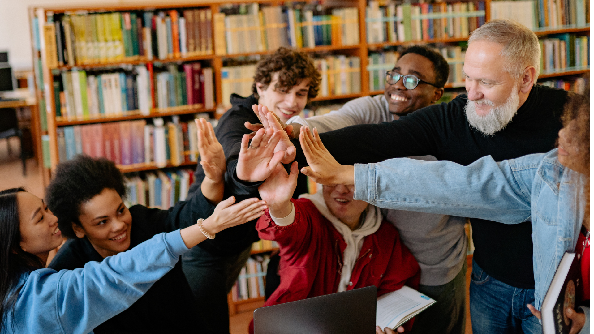 Group of coworkers high-fiving in a library, smiling around a laptop and bookshelves.