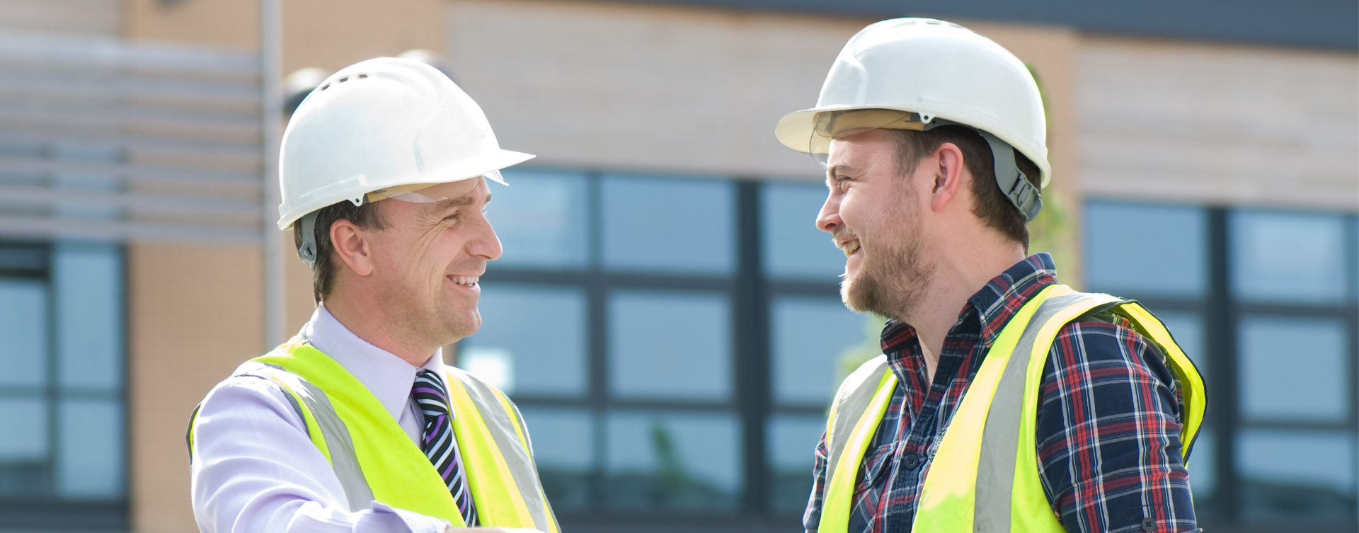 Two construction workers in hard hats and safety vests talking outside a building