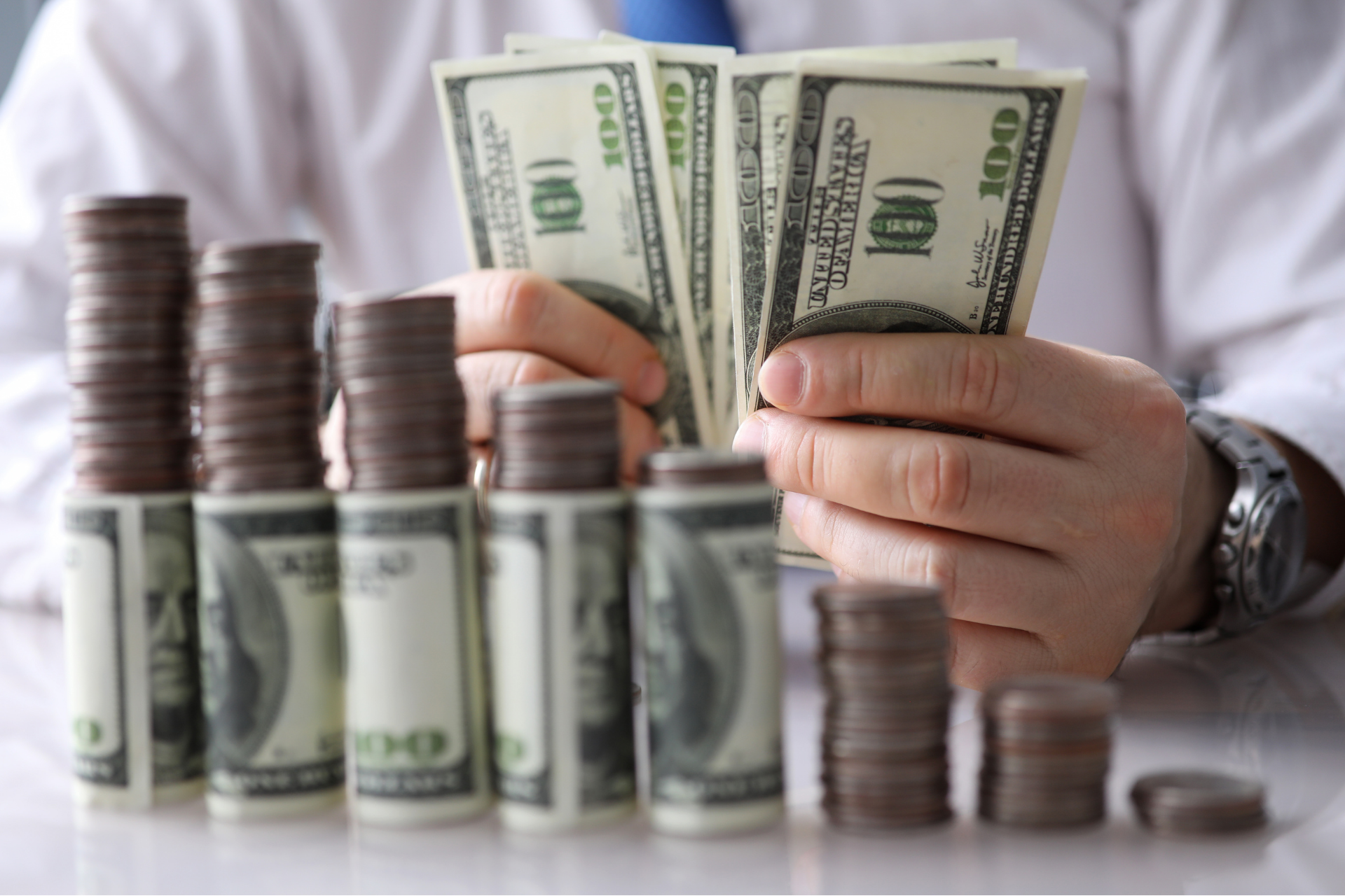 Hands counting U.S. dollar bills beside stacks of coins and cash bundles on a table