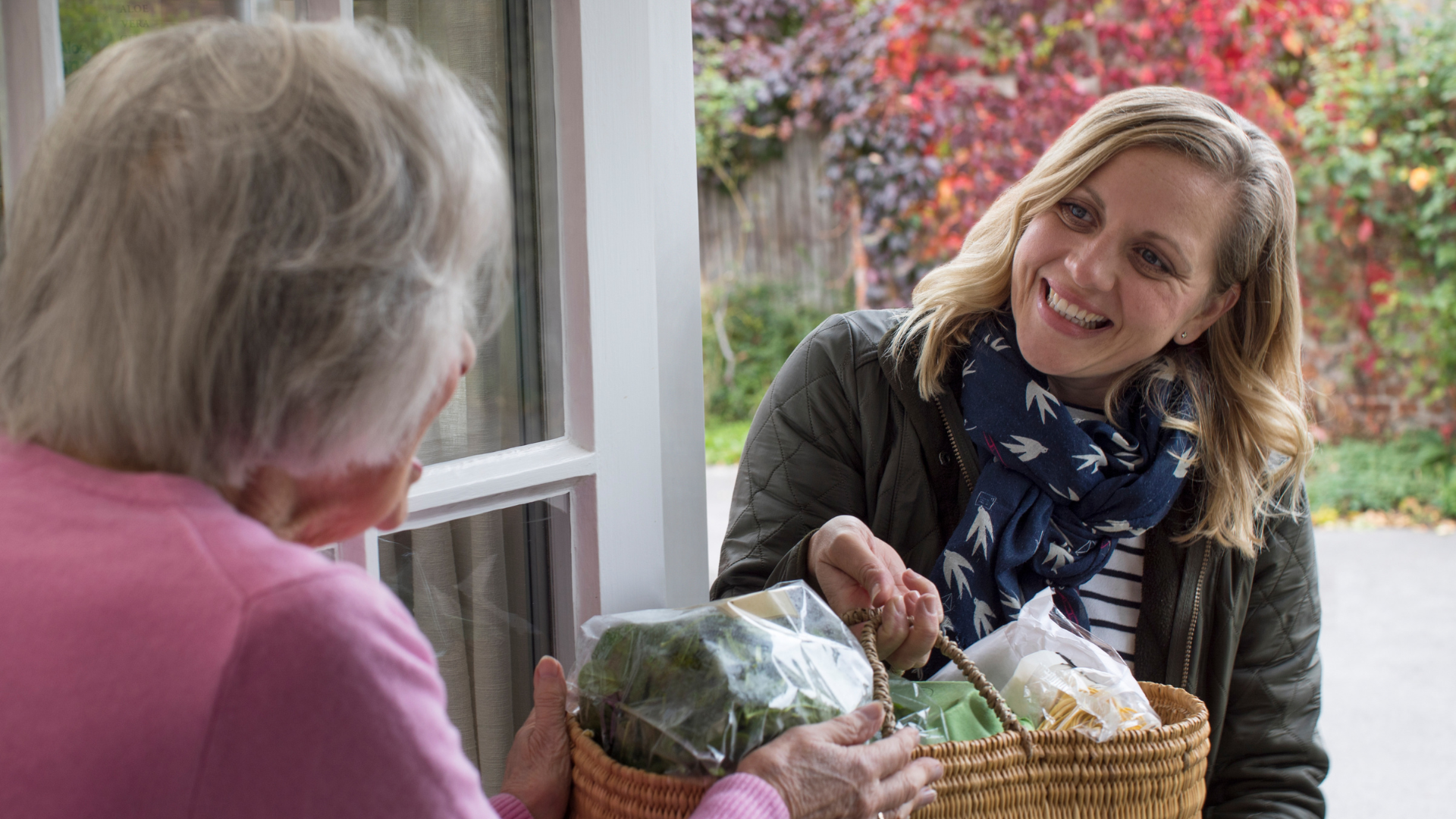 Two people chatting by a window, one holding a basket of flowers and greenery.
