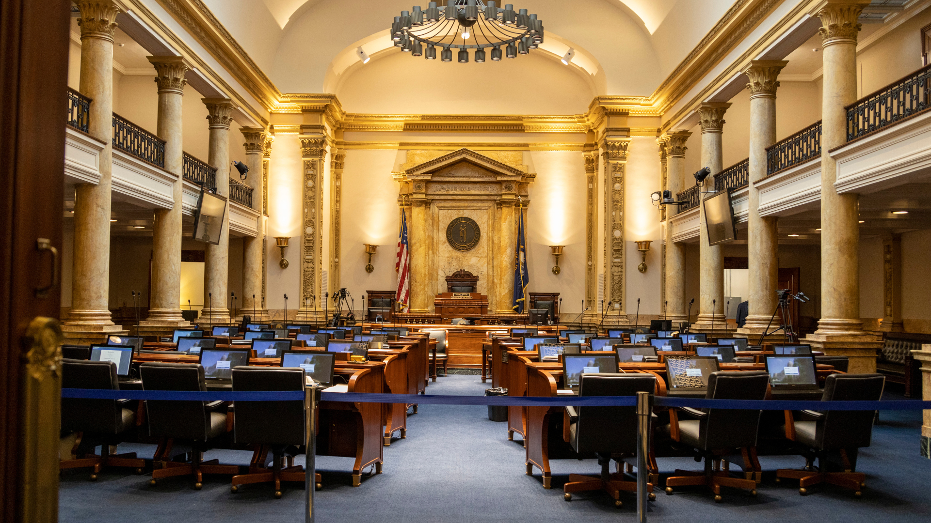 Elegant courtroom with rows of wooden desks, blue chairs, and a central bench beneath a chandelier.