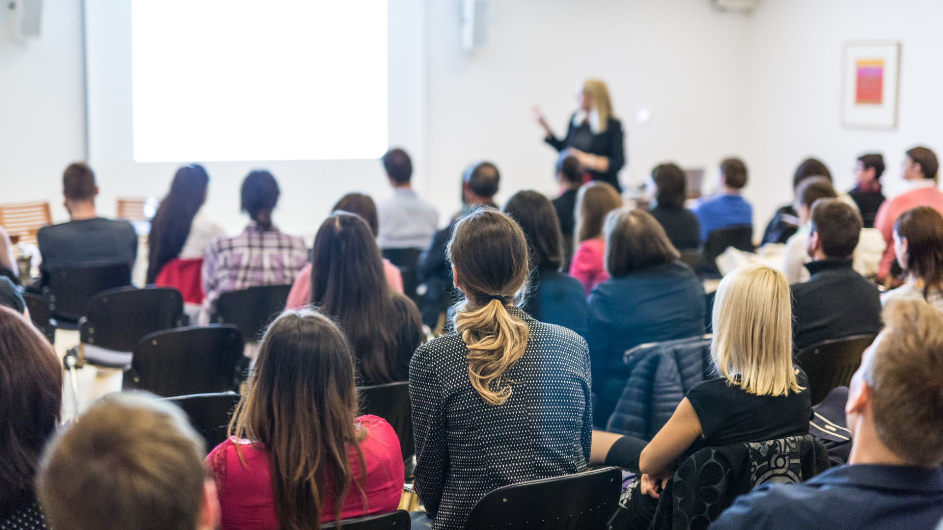 Audience seated in a conference room, watching a speaker at the front.