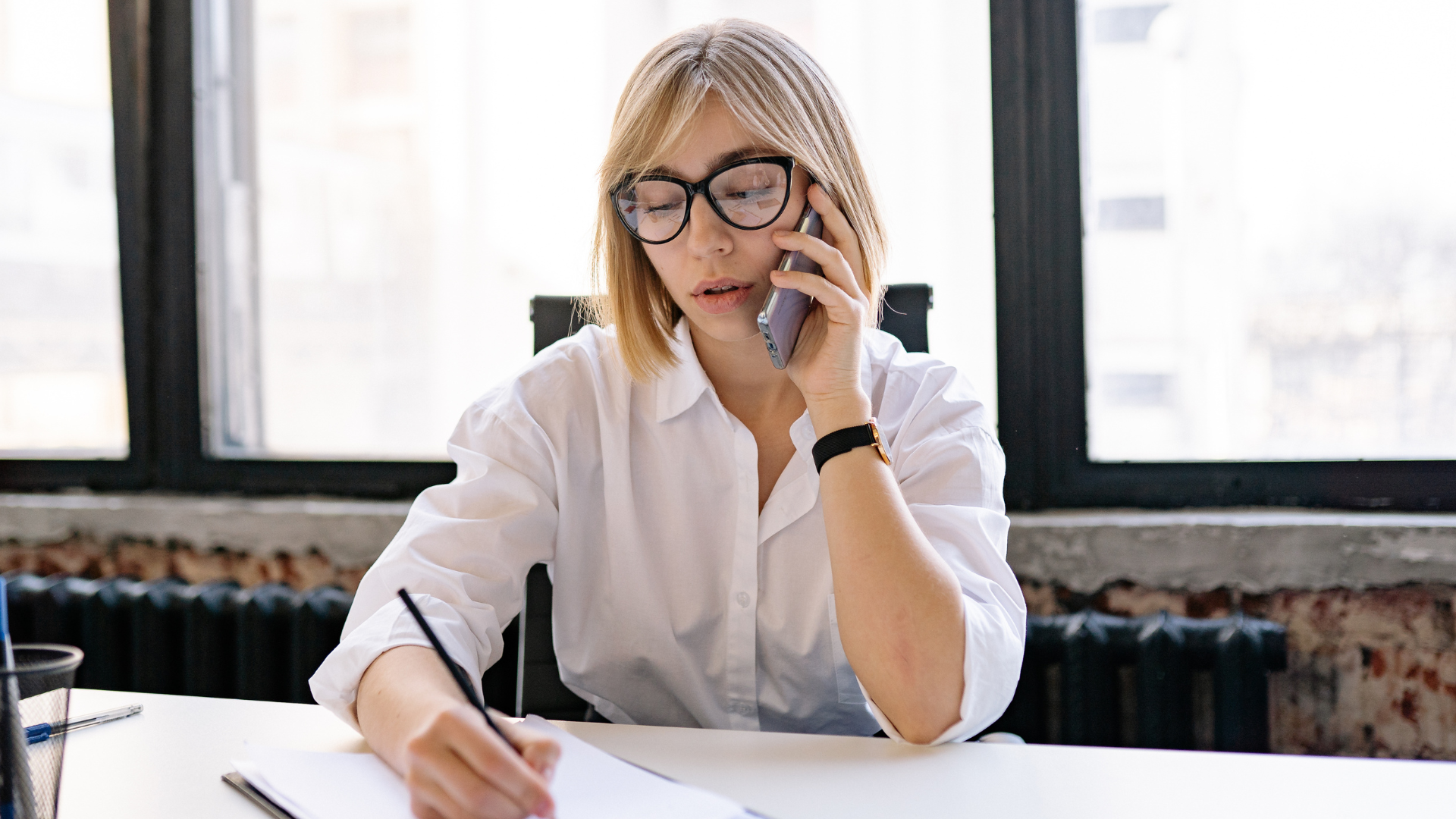 Woman in glasses writing at a desk and speaking on a phone in a bright office setting