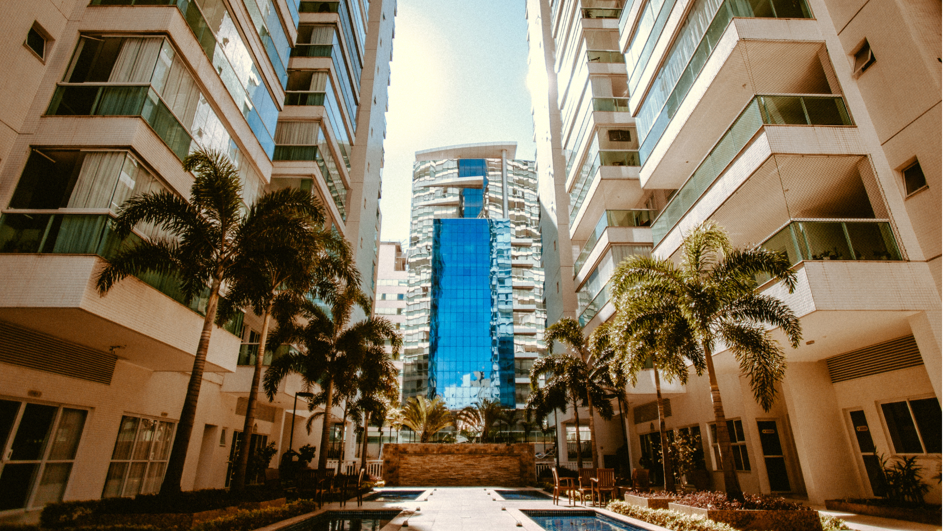 Modern white apartment buildings with palm trees framing a sunlit central walkway and a blue tower in the distance