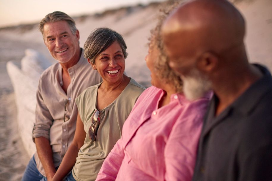 Three friends chatting on a beach at sunset, smiling and seated in a row.
