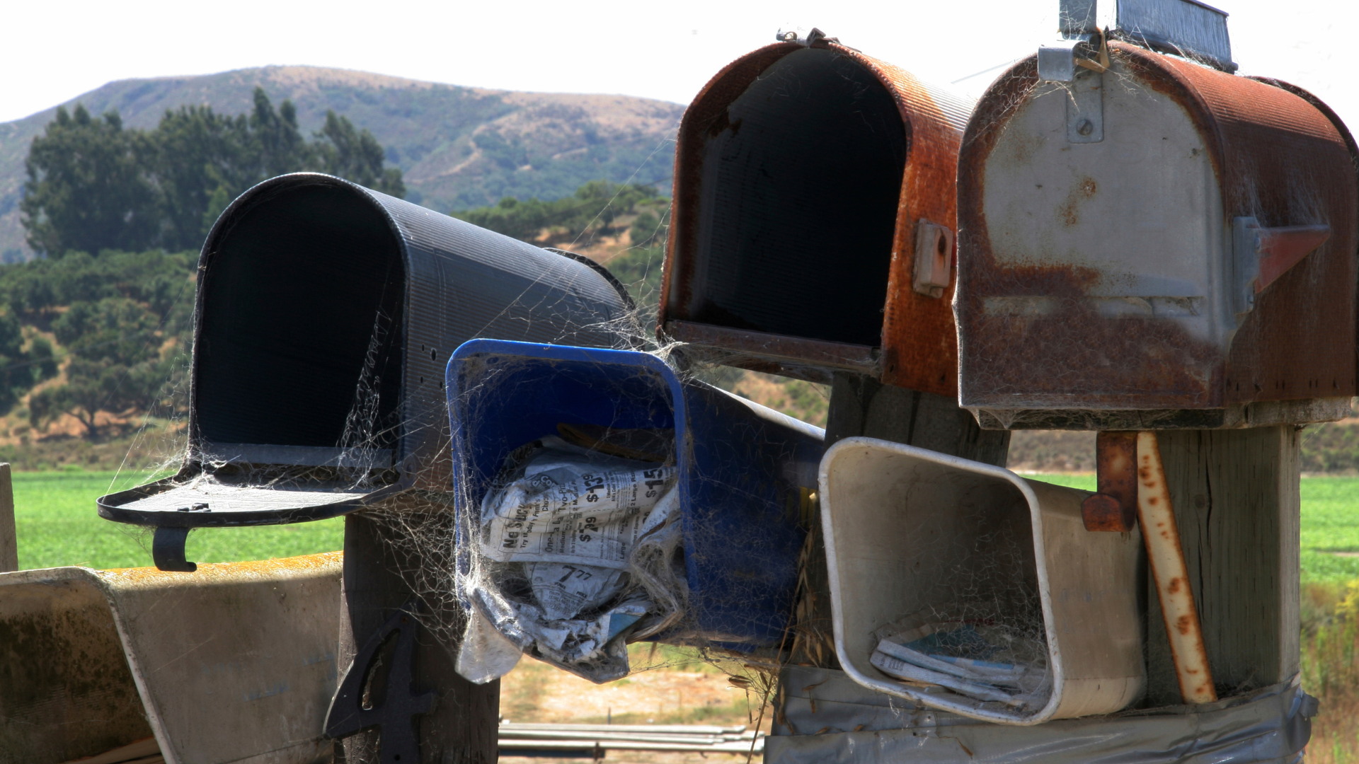 Three old metal mailboxes, one blue and two rusted, standing outdoors with hills in the background