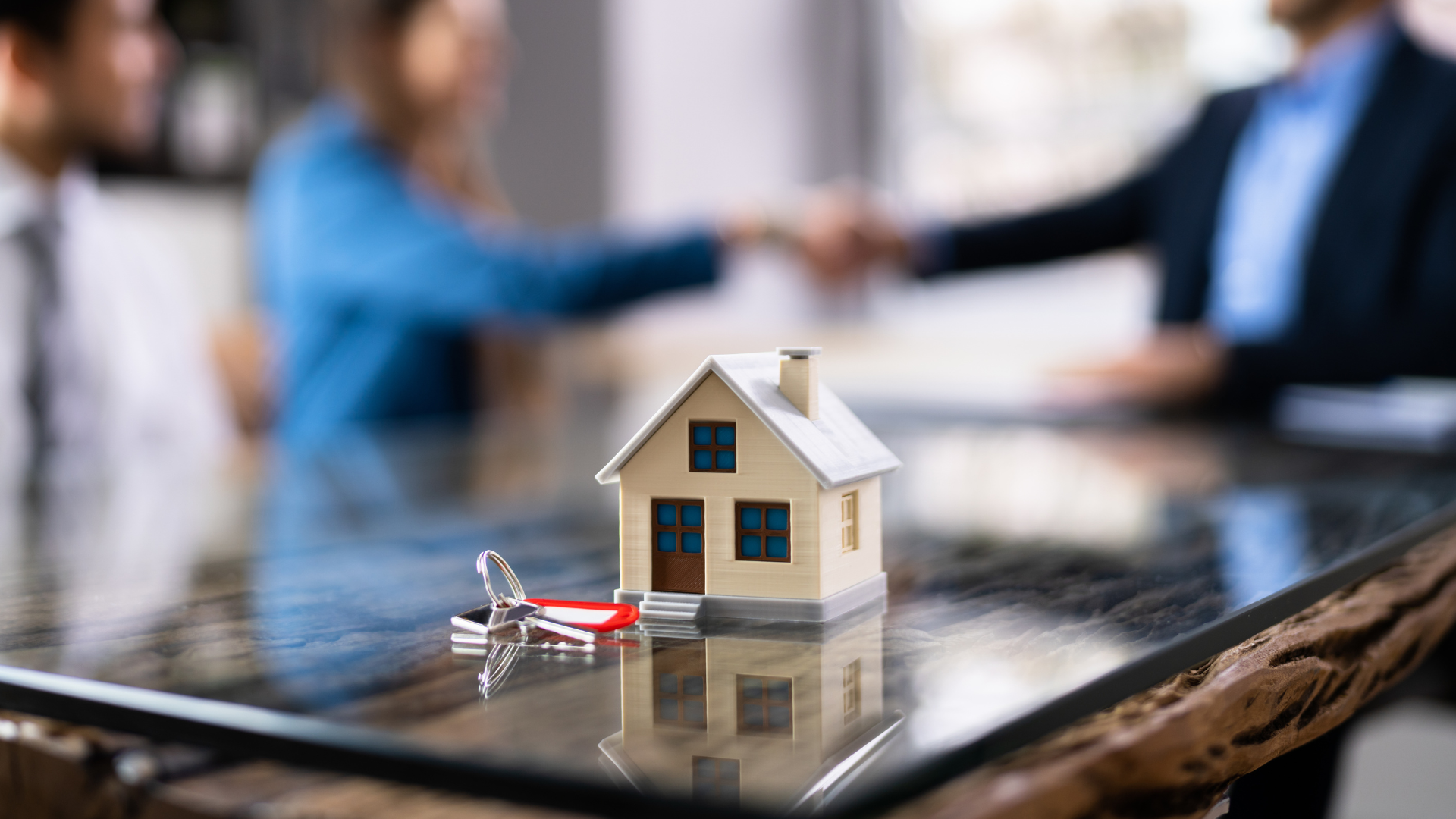 Miniature house model with keys on a tablet, with blurred people shaking hands in the background