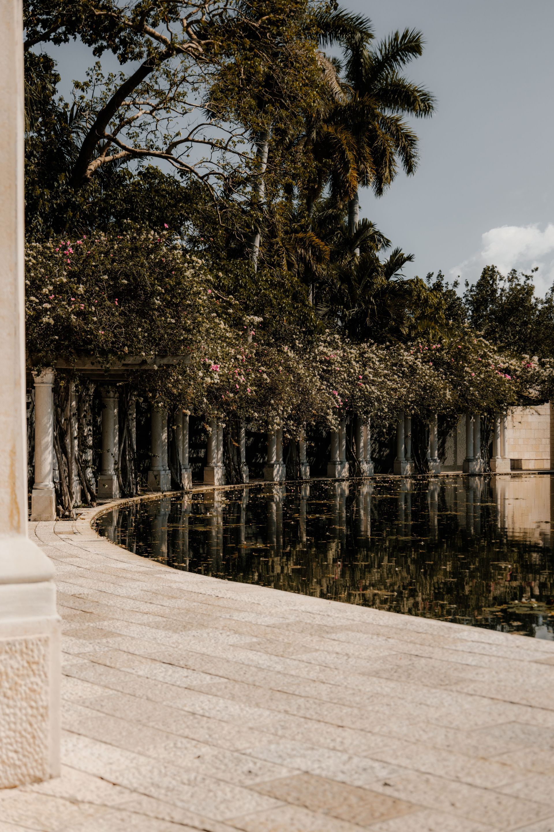 Curved stone walkway beside a calm reflecting pool, lined with trees and a white columned wall