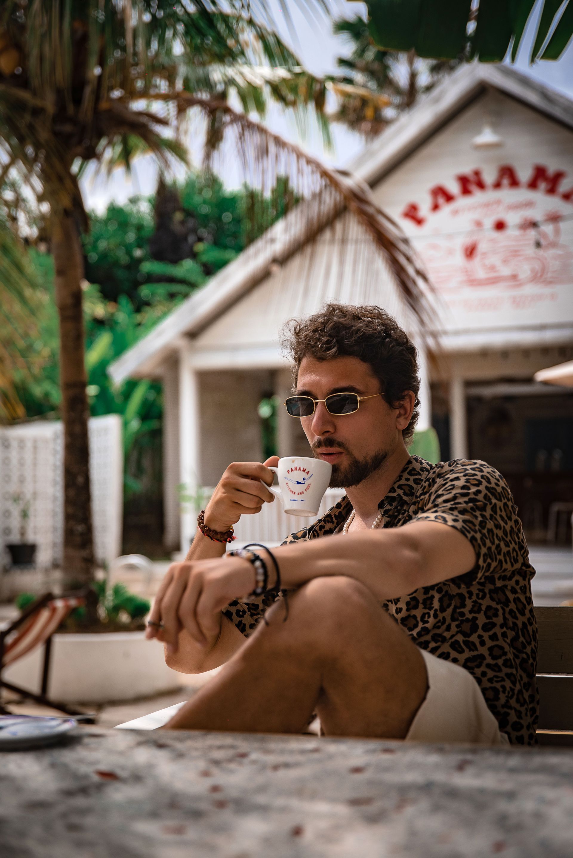 Man sitting outdoors in a leopard-print shirt and sunglasses, holding a mug in front of a white tropical hut