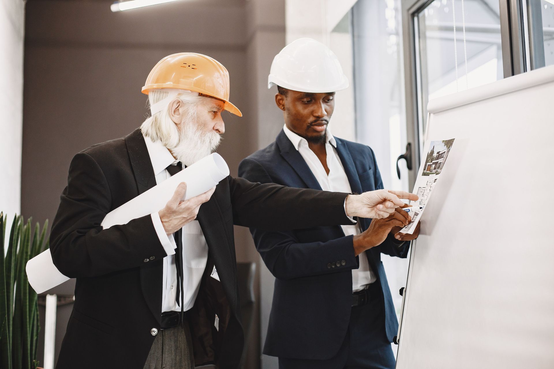 Two engineers in hard hats reviewing plans and pointing at a whiteboard indoors