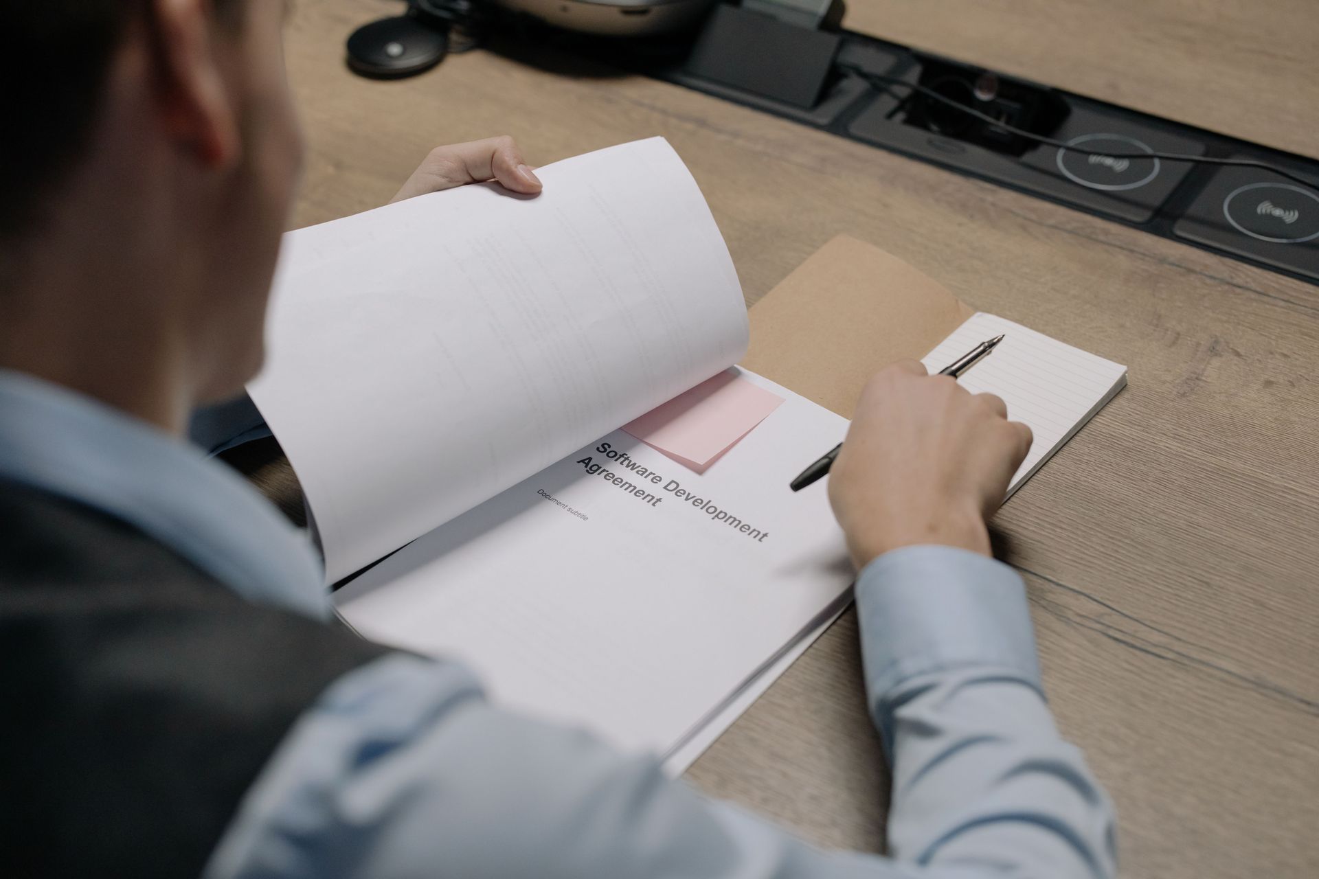 Person reviewing documents on a desk, writing in a notebook with a pen.