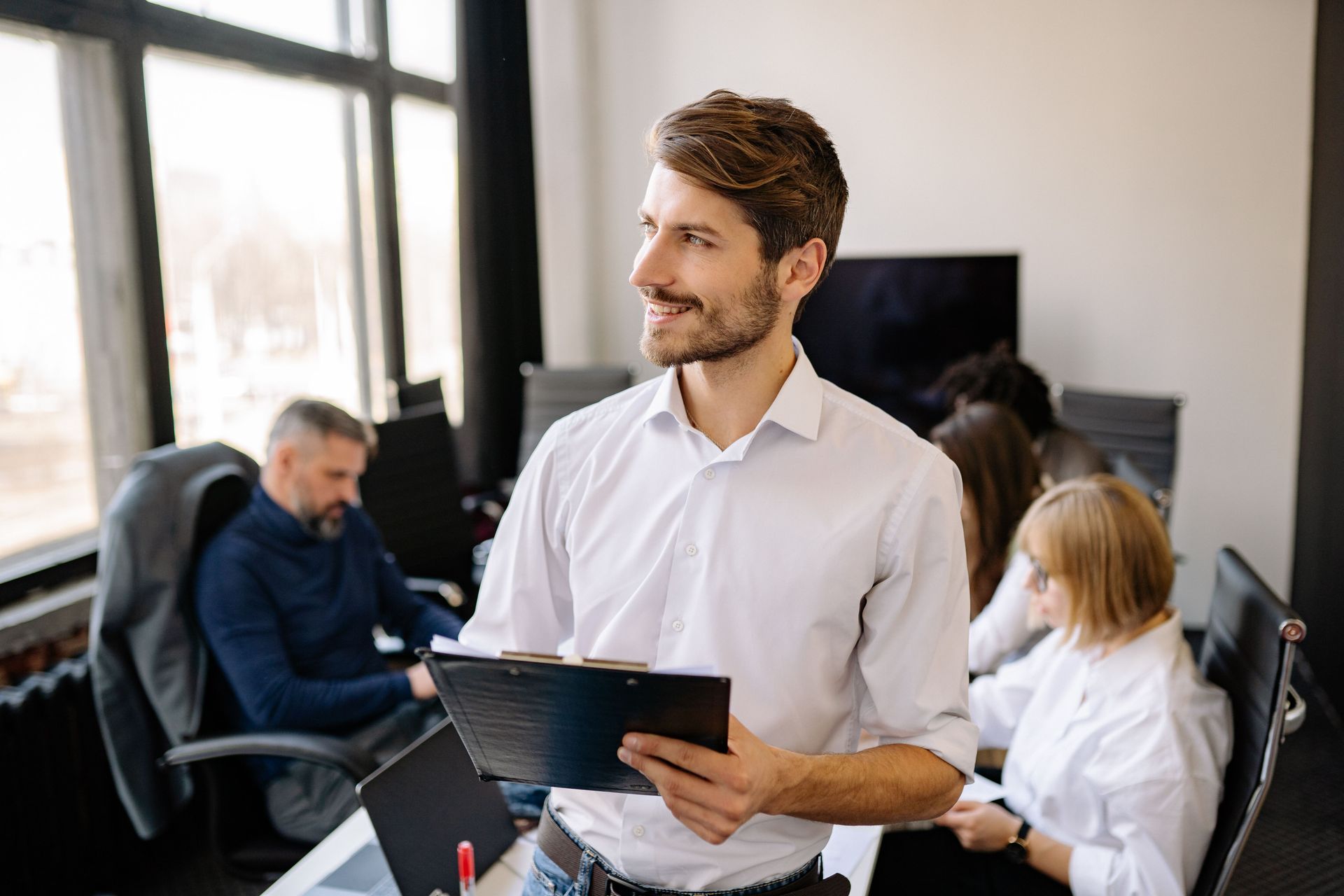 Man in white shirt holding a tablet in a modern office with coworkers in the background