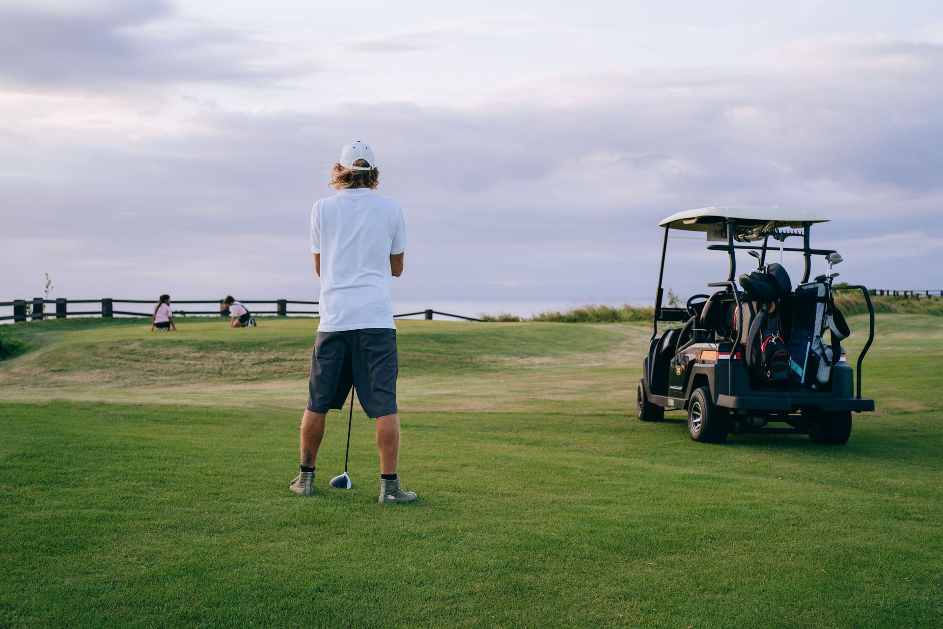Golfer on a green fairway near a golf cart at dusk, facing the open course.