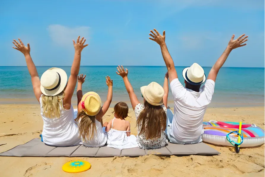 Family sitting on a beach, raising their arms toward the ocean under a bright blue sky