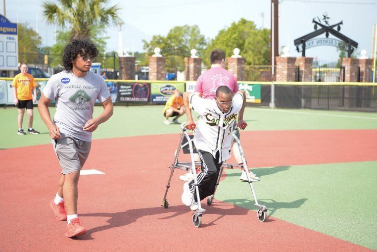A man in a gray shirt is walking a man in a walker on a baseball field.