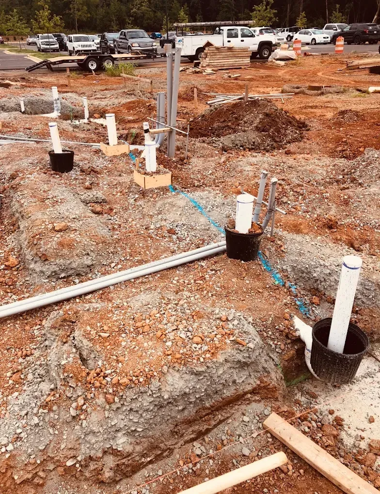 Construction site with exposed pipes, trenches, and dirt; cars parked in background.