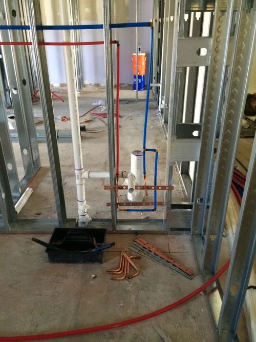 Exposed wall framing with plumbing pipes, a black toolbox, and coiled red cable on a concrete floor.